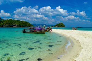 A serene seaside view of Krabi, Thailand, with a stretch of golden sand bordered by the vibrant Andaman Sea. In the foreground, a colorful longtail boat rests gently on the shore, while other traditional boats drift lazily in the turquoise waters. The backdrop showcases stunning islands and rocky formations rising from the sea, creating an idyllic landscape that blends crystal-clear water with the natural beauty of Thailand’s tropical coastline. The scene captures the essence of a peaceful day by the beach, perfect for relaxation and adventure.